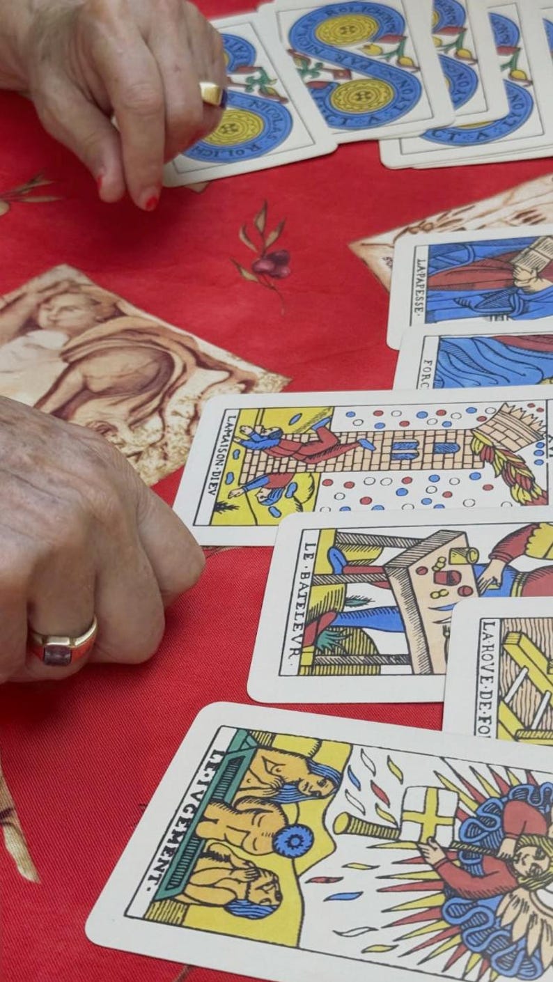 May include: A selection of Tarot cards with vibrant illustrations are arranged on a red surface. The cards display images and French text. A person wearing a gold ring is holding the cards.