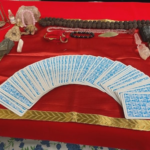 May include: A collection of spiritual items arranged on a red surface. A deck of tarot cards with blue and white designs is fanned out. Other items include crystals, beads, and small decorative objects. The scene is framed with a gold-trimmed border.