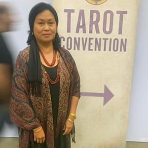 May include: A woman stands in front of a banner that reads "TAROT CONVENTION." The banner has a purple arrow pointing right and a circular image of a crowned figure. The woman wears a black head covering, a patterned shawl, and red beaded necklaces.