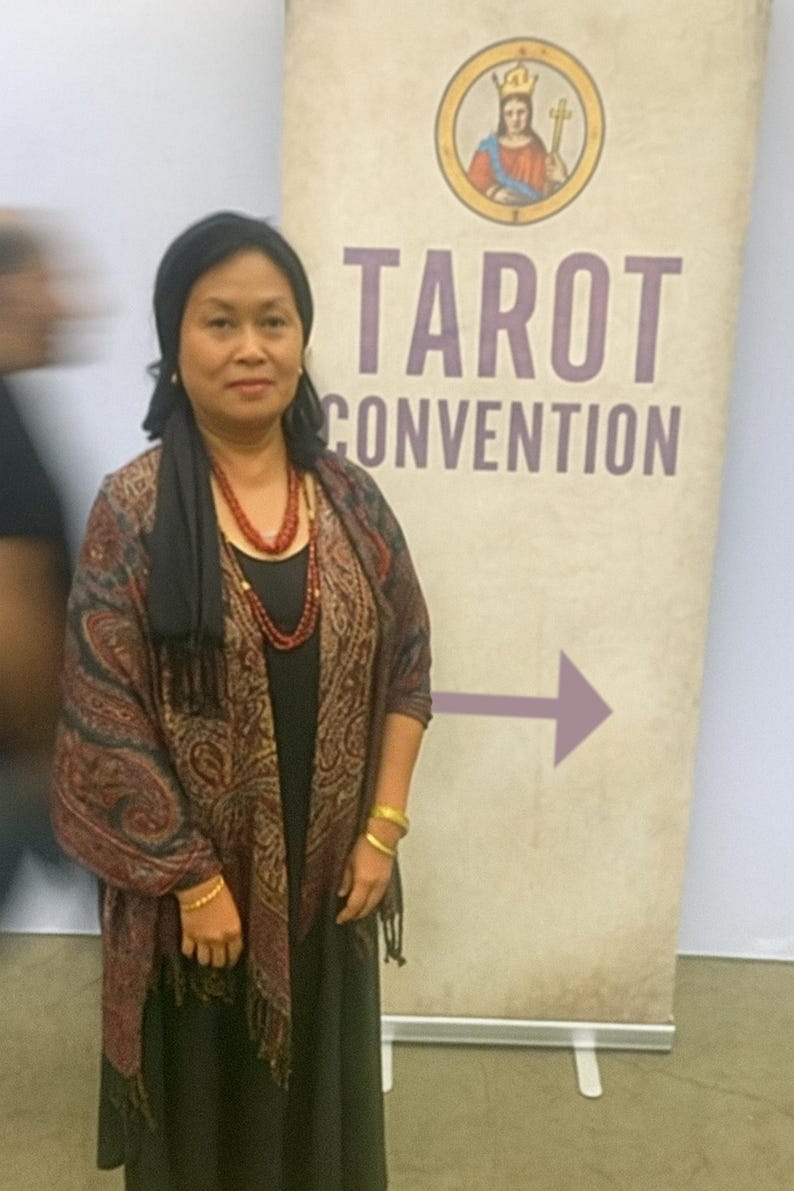 May include: A woman stands in front of a banner that reads "TAROT CONVENTION." The banner has a purple arrow pointing right and a circular image of a crowned figure. The woman wears a black head covering, a patterned shawl, and red beaded necklaces.