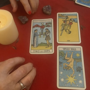 May include: Tarot card reading with four cards visible: "Two of Cups," "The Star," and another card. The cards are on a red surface, with crystals and a lit candle. The cards have illustrations and text.
