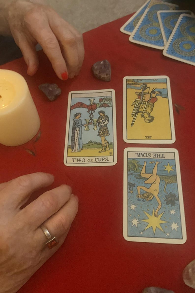 May include: Tarot card reading with four cards visible: "Two of Cups," "The Star," and another card. The cards are on a red surface, with crystals and a lit candle. The cards have illustrations and text.
