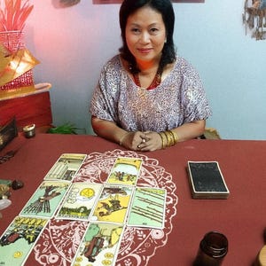 May include: A woman sits at a table with tarot cards laid out in a spread. The cards are colorful with various illustrations. A deck of cards, a small brown bottle, and crystals are also on the red table. The woman wears a patterned top and jewelry.