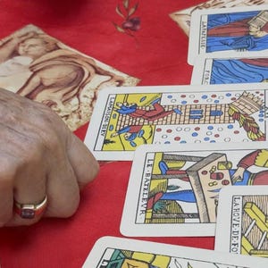 May include: Tarot cards with colorful illustrations are displayed on a red surface. The cards feature images and French text. The cards are being held by a person wearing a gold ring.