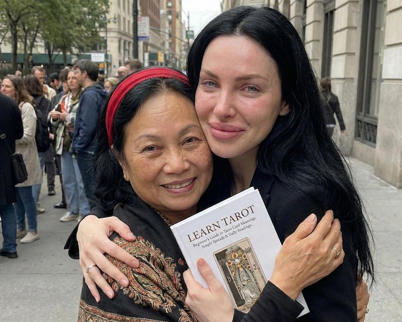 May include: Two women embracing outdoors, one holding a book titled "LEARN TAROT." The book cover features a tarot card illustration. The women are smiling, with a crowd and buildings in the background.