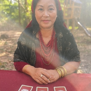 May include: A woman wearing a red headscarf and jewelry sits at a red table with tarot cards. She is wearing a red dress and a black shawl. The background is a blurred outdoor scene with trees and a small wooden structure.