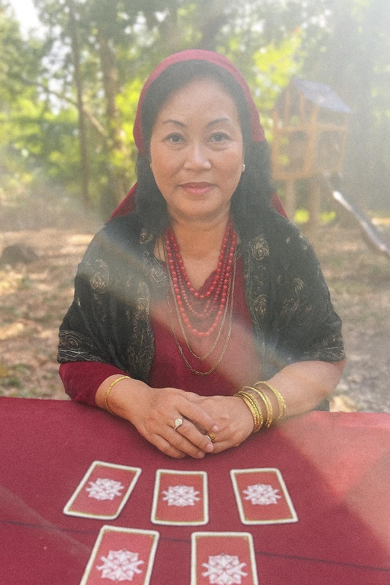 May include: A woman wearing a red headscarf and jewelry sits at a red table with tarot cards. She is wearing a red dress and a black shawl. The background is a blurred outdoor scene with trees and a small wooden structure.