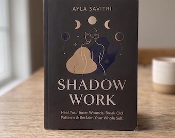 Puede incluir: Un libro negro titulado Shadow Work de Ayla Savitri. La portada presenta una ilustración lineal de una persona, fases lunares y formas abstractas en beige y azul oscuro. El subtítulo dice: Heal Your Inner Wounds, Break Old Patterns & Reclaim Your Whole Self.