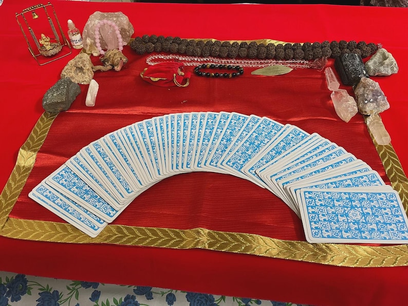 May include: A display of spiritual items on a red cloth. A deck of tarot cards with blue and white patterns is fanned out. Other items include crystals, beads, and small decorative objects. The scene is framed with a gold-trimmed border.
