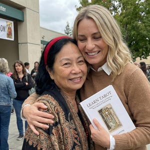 May include: Two women embracing outdoors, one holding a book titled "Learn Tarot." The book's cover features an illustration. The women are smiling. A banner in the background reads "Convention."