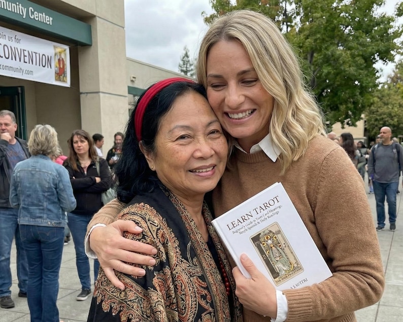 May include: Two women embracing outdoors, one holding a book titled "Learn Tarot." The book's cover features an illustration. The women are smiling. A banner in the background reads "Convention."