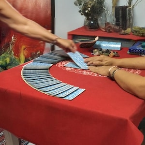 May include: A tarot card reading in progress. A deck of blue-backed tarot cards is fanned out on a red tablecloth. Two sets of hands are visible, one shuffling and dealing cards, the other resting on the table. The scene is set in a room with a red painting.