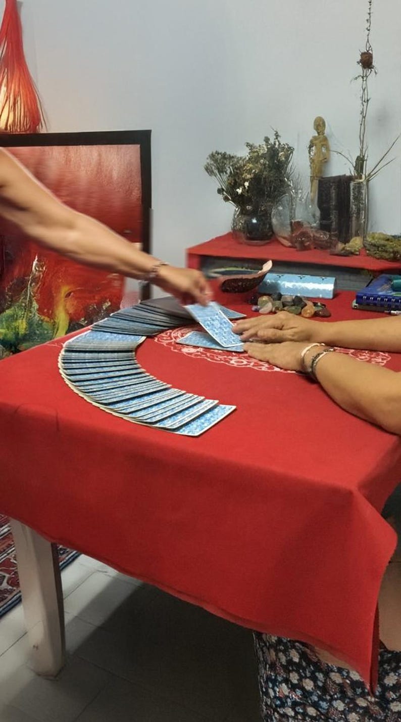 May include: A tarot card reading in progress. A deck of blue-backed tarot cards is fanned out on a red tablecloth. Two sets of hands are visible, one shuffling and dealing cards, the other resting on the table. The scene is set in a room with a red painting.