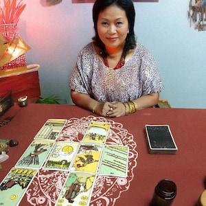 May include: A woman sits at a table with tarot cards laid out in a spread. The cards are colorful with various illustrations. A deck of cards, a small brown bottle, and crystals are also on the red table. The woman wears a patterned top and jewelry.