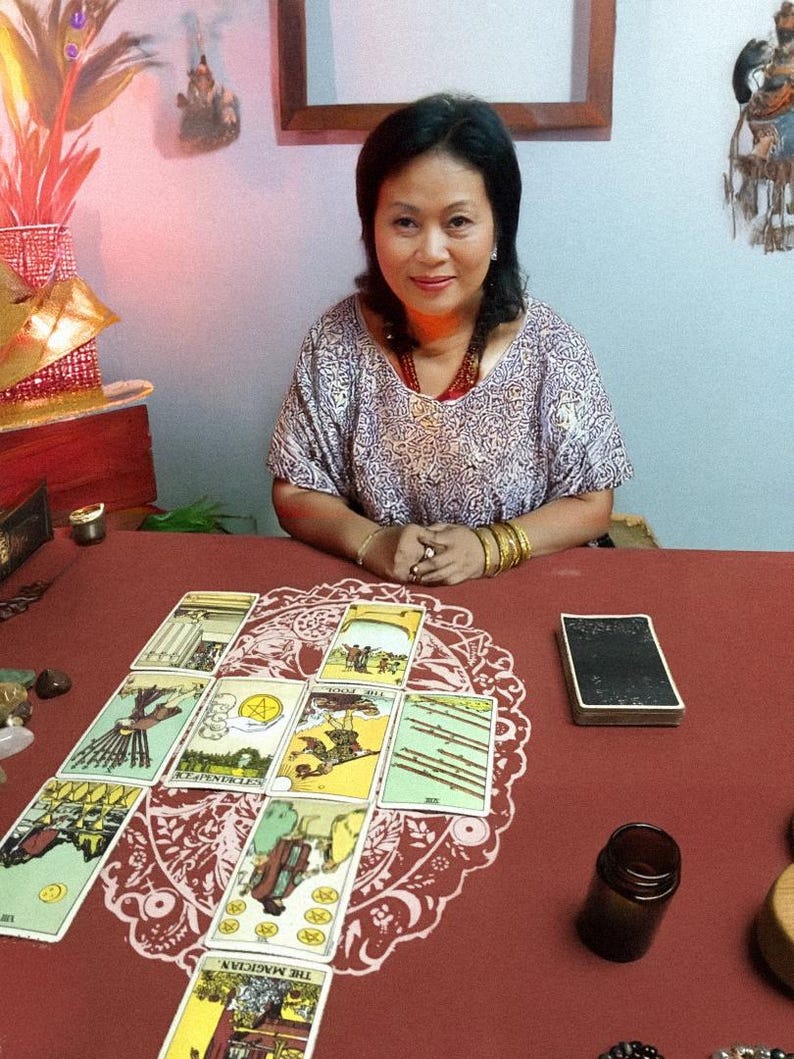 May include: A woman sits at a table with tarot cards laid out in a spread. The cards are colorful with various illustrations. A deck of cards, a small brown bottle, and crystals are also on the red table. The woman wears a patterned top and jewelry.