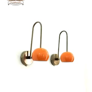 Könnte beinhalten: Zwei Wandleuchten mit orangefarbenen Kugelschirmen und gebogenen Metallarmen. Die Leuchten haben eine messingfarbene Basis und ein minimalistisches Design, ideal, um jedem Raum einen Hauch von modernem Stil zu verleihen. Das Logo von The Eagle Crafts befindet sich oben links.