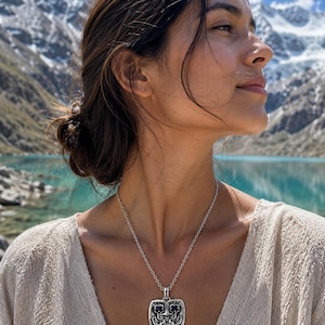 May include: A silver pendant necklace with a square, detailed pendant featuring a stylized face design. The necklace is worn against a light-colored top, with a scenic mountain and lake backdrop.