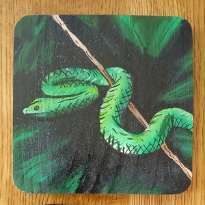 May include: A square coaster featuring a painted illustration of a bright green snake coiled around a thin brown branch. The background consists of dark green foliage with visible brushstrokes, creating a dense, shadowed forest environment.