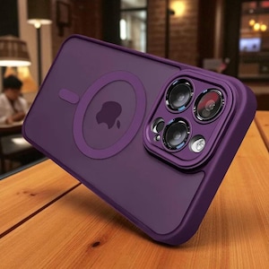 May include: A purple phone case with a camera lens protector. The case has a circular magnetic ring and an Apple logo. The case is on a wooden surface.