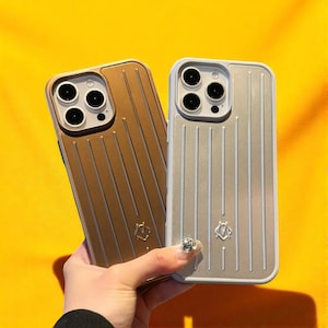 May include: Two phone cases with a vertical striped design. One case is gold, the other is silver. Both cases have a camera cutout and a small emblem at the bottom. The background is a bright yellow.