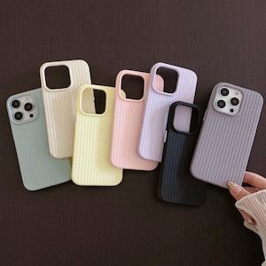 May include: A collection of colorful phone cases in various pastel shades, including light blue, cream, yellow, pink, lavender, black, and gray. Each case features a ribbed texture and cutouts for the camera and buttons. The cases are arranged on a dark brown surface.