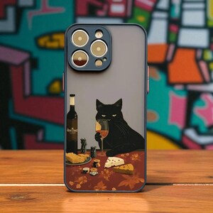 Puede incluir: Funda de teléfono con un gato negro bebiendo vino, una botella de vino y queso sobre una mesa. La funda tiene un borde azul y un fondo gris. La mesa tiene un estampado floral rojo.