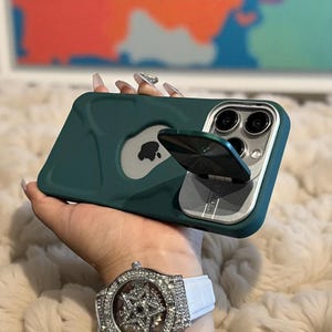 Op de afbeelding: Donkergroene telefoonhoes met een zilveren Apple-logo en een zilveren ringhouder. De hoes heeft een metallic zilveren rand rond de cameralenzen. De ringhouder is open en schuin.