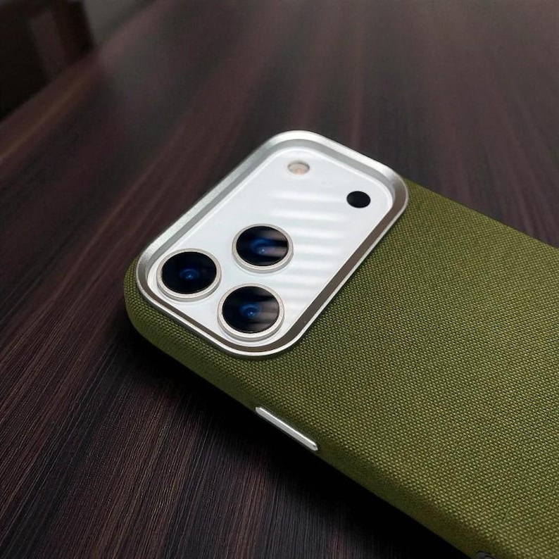 May include: An olive green phone case with a silver camera lens protector. The case has a textured fabric appearance. The camera lens protector has three circular lenses and a small black dot.