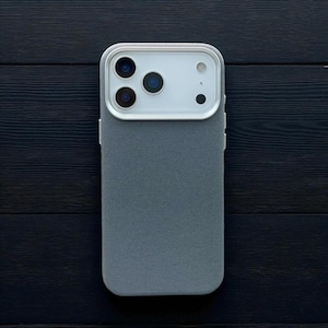 May include: A grey phone case with a textured fabric finish. The case features a white camera module with three lenses and a flash. The phone case is on a dark wood surface.