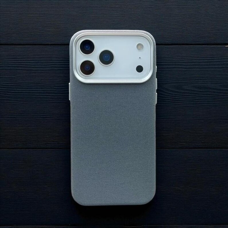 May include: A grey phone case with a textured fabric finish. The case features a white camera module with three lenses and a flash. The phone case is on a dark wood surface.