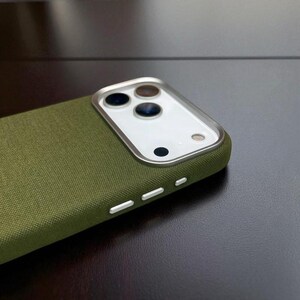 May include: Olive green phone case with a textured fabric finish. The case has a silver camera frame and white buttons. The camera area has three lenses and a flash.
