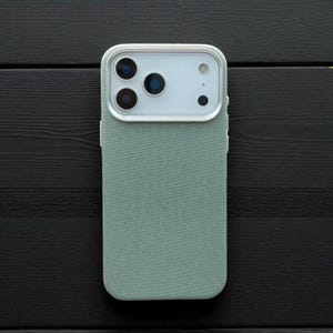 May include: A mint green phone case with a textured fabric finish. The case has a white camera module with three lenses and a flash. The phone case is shown on a black wooden surface.