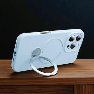 May include: A pale blue phone case featuring a silver ring holder. The case rests on a wooden surface against a dark backdrop. The ring holder is attached to the rear and functions as a stand.