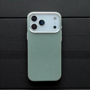 May include: A mint green phone case with a textured fabric finish. The case has a white camera module with three lenses and a flash. The phone case is shown on a black wooden surface.