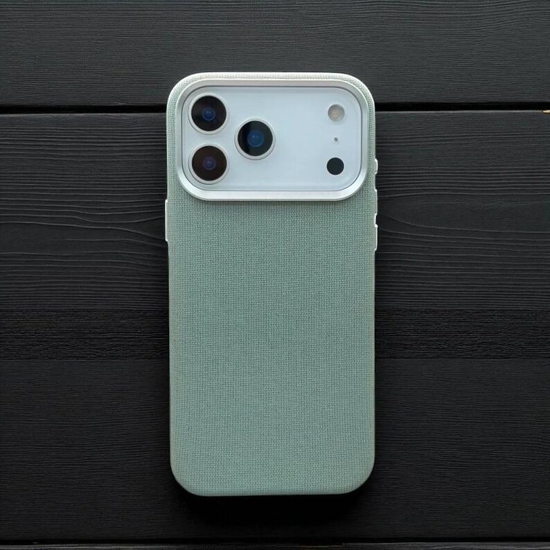 May include: A mint green phone case with a textured fabric finish. The case has a white camera module with three lenses and a flash. The phone case is shown on a black wooden surface.