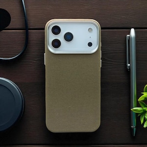 May include: A khaki phone case with a silver camera frame. The case is on a dark wooden surface, next to a silver pen and a black object. The phone case has three camera lenses and a flash.