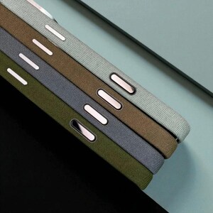 May include: A close-up shot of several mobile phone cases in shades of green, grey and brown. Each case has a textured fabric finish and metallic buttons. The cases are stacked at an angle, showing their side profiles.
