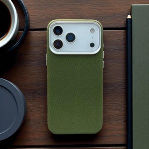 May include: Olive green phone case with a white camera module. The case has a textured fabric appearance. The phone case is on a wooden surface, next to a pencil, a notebook, and a cup of coffee.