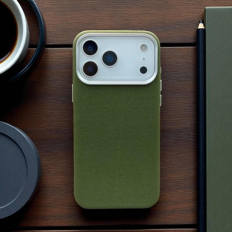 May include: Olive green phone case with a white camera module. The case has a textured fabric appearance. The phone case is on a wooden surface, next to a pencil, a notebook, and a cup of coffee.