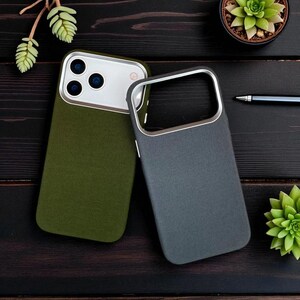May include: Two mobile phone cases, one olive green and one grey, with silver-coloured metal trim. The cases have a textured finish and are on a dark wooden surface, with a pen and small plants.