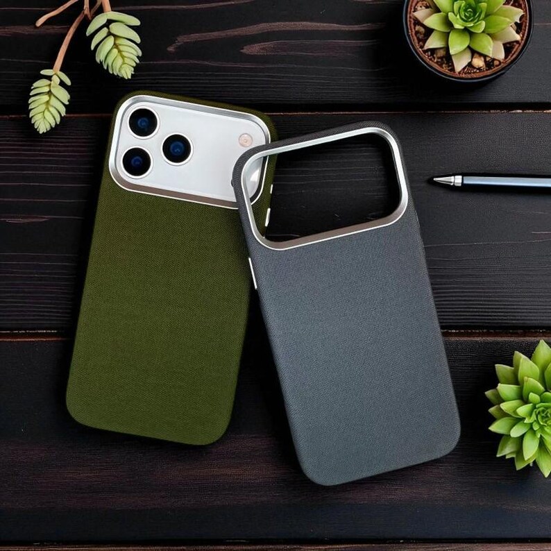 May include: Two mobile phone cases, one olive green and one grey, with silver-coloured metal trim. The cases have a textured finish and are on a dark wooden surface, with a pen and small plants.