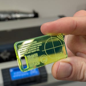 Puede incluir: Un dispositivo rectangular amarillo transparente con un borde verde y texto. El texto dice "FIELD COMPASS FOR WILDERNESS NAVIGATORS WITH ALIGN CLUSTER TO NEBULAE JOY CORP HQ." También se ve un código QR y una retícula de puntería.