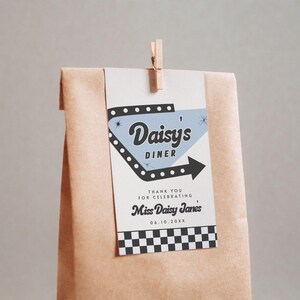 May include: A brown paper bag with a tag that reads "Daisy's Diner" in a retro font. The tag has a black arrow and a checkered pattern at the bottom. A wooden clip holds the tag in place.