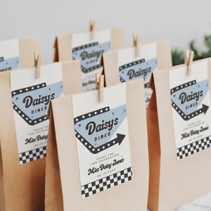 May include: Several kraft paper favor bags with tags that read "Daisy's Diner." The tags have a retro diner design in blue and black, with a black arrow. Each bag is clipped with a small wooden clothespin. The bags are for a celebration.