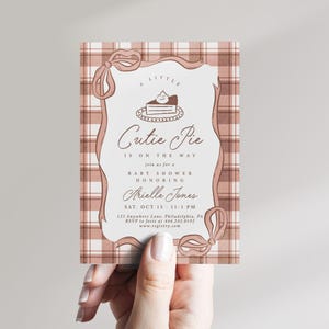 May include: A rectangular baby shower invitation card featuring a dusty rose and white plaid background. The center white panel is framed by a dusty rose ribbon border with bows and includes a pie illustration and text: A Little Cutie Pie is on the way.