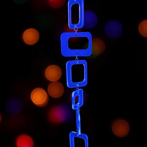 May include: A vertical chain of six translucent blue acrylic rectangular links connected by small silver metal jump rings. The links vary slightly in size and are arranged in a linear sequence, hanging from a thin black cord at the top.