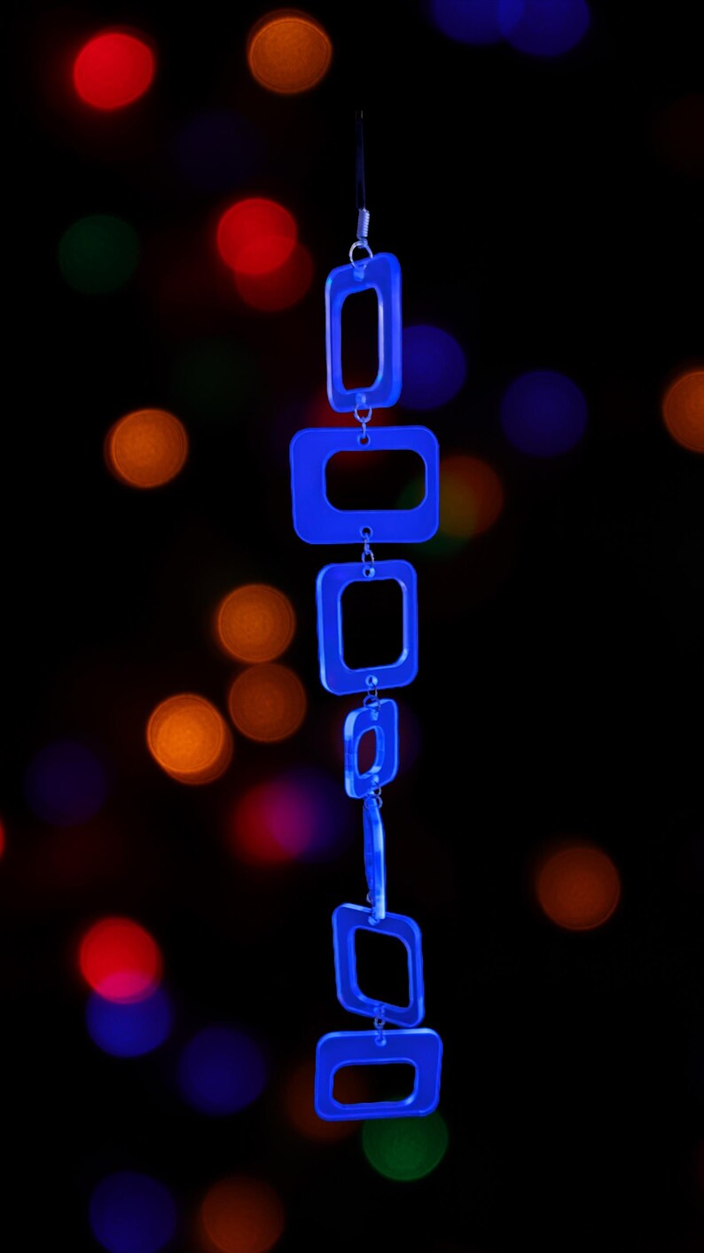 May include: A vertical chain of six translucent blue acrylic rectangular links connected by small silver metal jump rings. The links vary slightly in size and are arranged in a linear sequence, hanging from a thin black cord at the top.