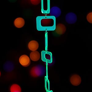May include: A vertical hanging mobile composed of five translucent neon green acrylic rectangular frames of varying sizes, linked together by small silver metal jump rings, suspended from a thin black cord against a dark background.