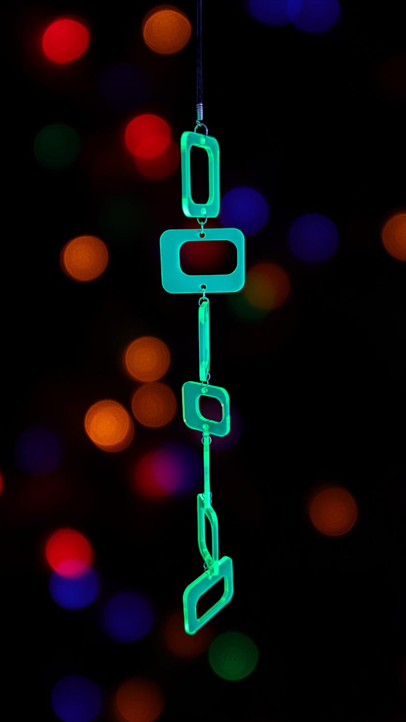May include: A vertical hanging mobile composed of five translucent neon green acrylic rectangular frames of varying sizes, linked together by small silver metal jump rings, suspended from a thin black cord against a dark background.