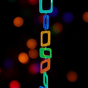 May include: A vertical hanging mobile composed of six translucent, neon-coloured square frames linked together. The frames are arranged in a sequence of light blue, dark blue, yellow, green, orange, and light blue, suspended by a thin black string.